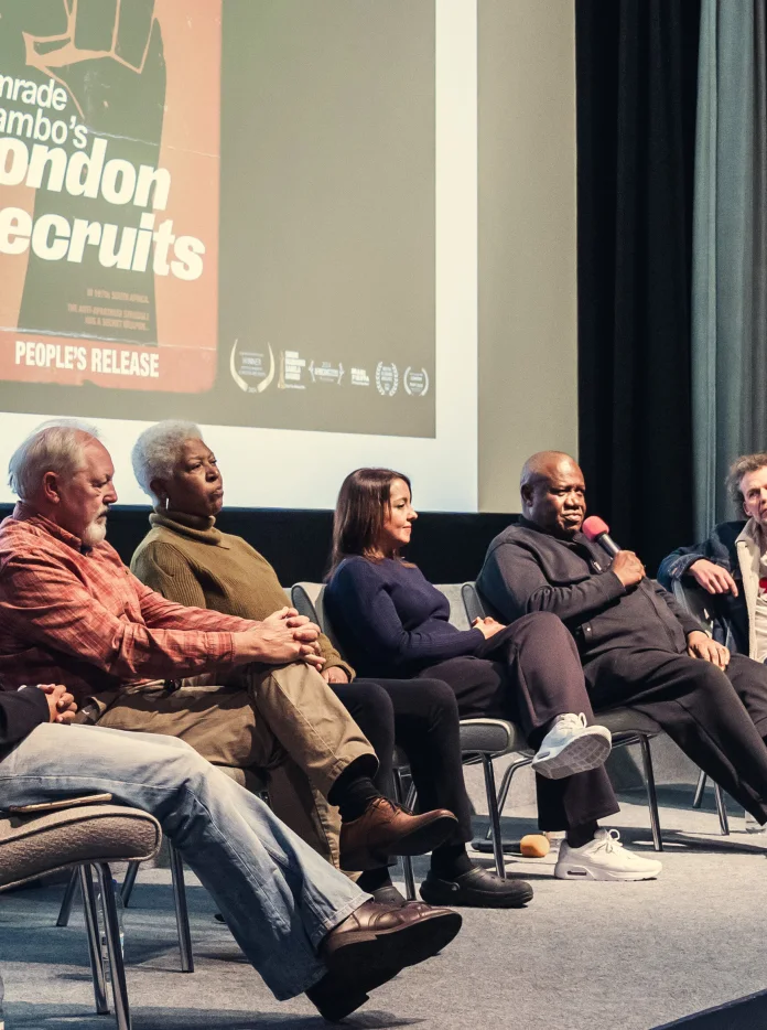 A panel of six speakers sits on stage in front of a projected film poster for Comrade Tambo’s London Recruits during a discussion event.